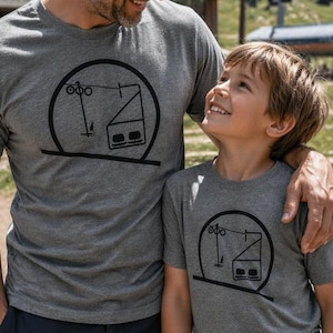 A father with his arm around his young son, both smiling at each other at an alpine resort during summer. They are wearing matching grey heather t-shirts with a black screen-printed minimalist circle graphic showing an alpine ski lift structure. In the background, a mountain ski lodge with a Big Sky sign is visible alongside a chairlift tower under a sunny sky.