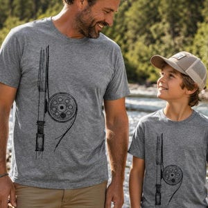 May include: Two grey t-shirts featuring a black fishing reel design. A man and a child are wearing the shirts and holding fishing rods. They are positioned near a river, with trees visible in the background.