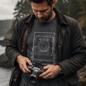 May include: A dark charcoal gray t-shirt featuring a white line-art graphic of a vintage large format camera. The shirt is worn under a dark brown waxed cotton jacket with a corduroy collar, paired with dark denim jeans and a silver wristwatch.