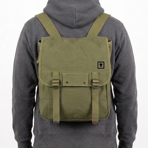 Peut inclure: Sac à dos en toile vert olive avec des bretelles réglables et une fermeture à rabat. Le sac à dos comporte deux fermetures à boucle et un petit logo carré sur le devant. Le sac est porté sur le dos.