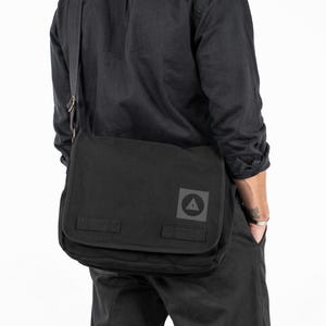 Puede incluir: Bolso mensajero negro con cierre de solapa y correa de hombro ajustable. El bolso presenta un parche cuadrado gris con un logotipo de triángulo. El bolso está hecho de un material duradero.