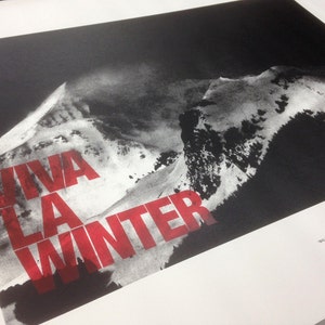 May include: Black and white print of a mountain range with the words "VIVA LA WINTER" in red block letters. The print has a stark contrast, with the mountain range appearing in shades of gray against a dark background.
