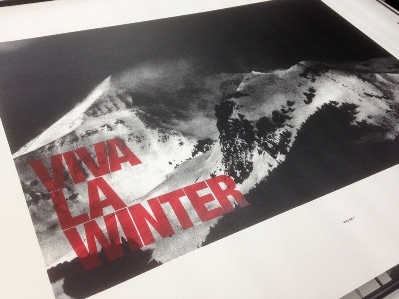 May include: Black and white print of a mountain range with the words "VIVA LA WINTER" in red block letters. The print has a stark contrast, with the mountain range appearing in shades of gray against a dark background.