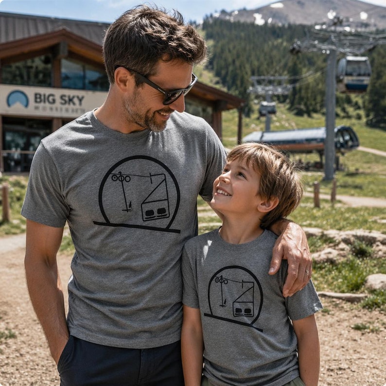 A father with his arm around his young son, both smiling at each other at an alpine resort during summer. They are wearing matching grey heather t-shirts with a black screen-printed minimalist circle graphic showing an alpine ski lift structure. In the background, a mountain ski lodge with a Big Sky sign is visible alongside a chairlift tower under a sunny sky.