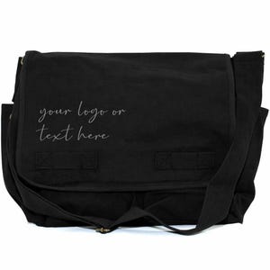 May include: Black messenger bag with a flap closure and adjustable shoulder strap. The bag has two front pockets and the text "your logo or text here" in a script font.