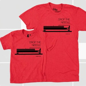 Flat lay product display of a matching father and child t-shirt set in vibrant heather red. Both shirts feature a bold, clean black industrial graphic of a vintage turntable profile with the typography "DROP THE NEEDLE" printed on the chest.