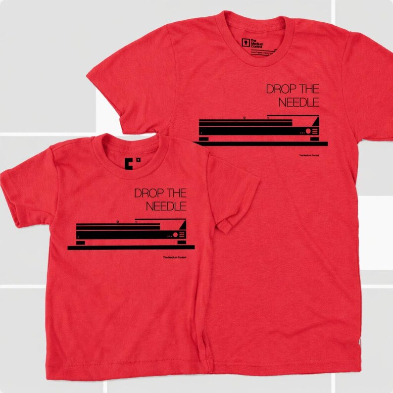 Flat lay product display of a matching father and child t-shirt set in vibrant heather red. Both shirts feature a bold, clean black industrial graphic of a vintage turntable profile with the typography "DROP THE NEEDLE" printed on the chest.