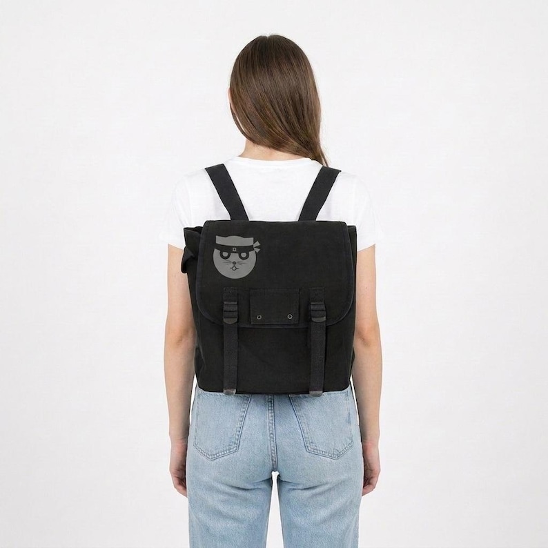 Back view of a person wearing a black canvas Ninja Watson backpack. The minimalist tech bag features a grey ninja cat graphic on the flap, showing the adjustable straps and unisex fit against a clean white background.