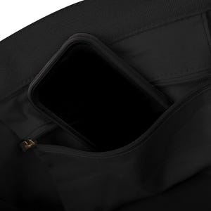 May include: Close-up of a black tote bag with a zipped pocket. A black smartphone is partially visible inside the bag's open pocket. The bag appears to be made of a durable fabric.