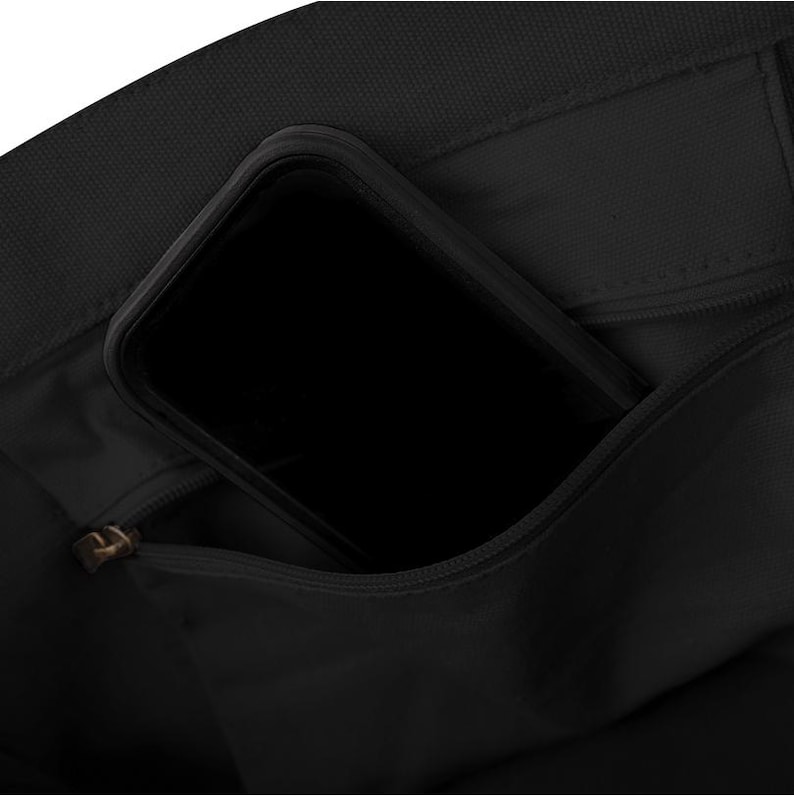 May include: Close-up of a black tote bag with a zipped pocket. A black smartphone is partially visible inside the bag's open pocket. The bag appears to be made of a durable fabric.