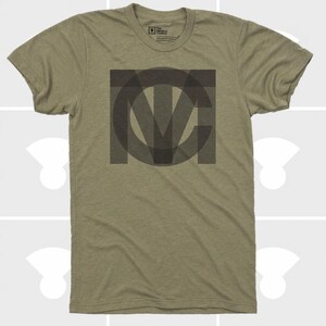 Pu&ograve; includere: T-shirt verde oliva con un design geometrico grigio scuro al centro. Il design presenta lettere sovrapposte, forse un'iniziale o un logo. La maglietta ha maniche corte e girocollo.
