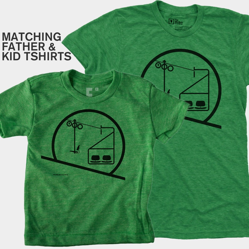 May include: Two green heathered t-shirts featuring a black graphic of a ski lift inside a circle. The text MATCHING FATHER & KID TSHIRTS is visible. The shirts have short sleeves and a crew neckline, displaying a consistent design on both garments.