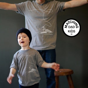 May include: Two gray t-shirts with matching white line art of a fishing scene. One shirt is worn by an adult, the other by a child. The shirts are part of a matching set. A black and white circular logo reads "Matching Dad + Kids".