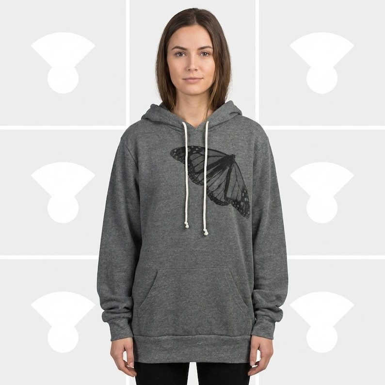 A woman wearing a heather grey Sponge Fleece pullover hoodie featuring a hand-printed butterfly graphic in black ink. The design is positioned as a minimalist, asymmetrical accent on the upper left chest, featuring a highly detailed, symmetrical scientific illustration of a butterfly.