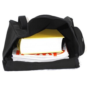 May include: Black canvas tote bag with a shoulder strap, filled with a stack of magazines and a yellow book. The bag has a side pocket with a button closure. The interior of the bag is visible.