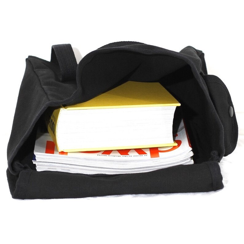 May include: Black canvas tote bag with a shoulder strap, filled with a stack of magazines and a yellow book. The bag has a side pocket with a button closure. The interior of the bag is visible.