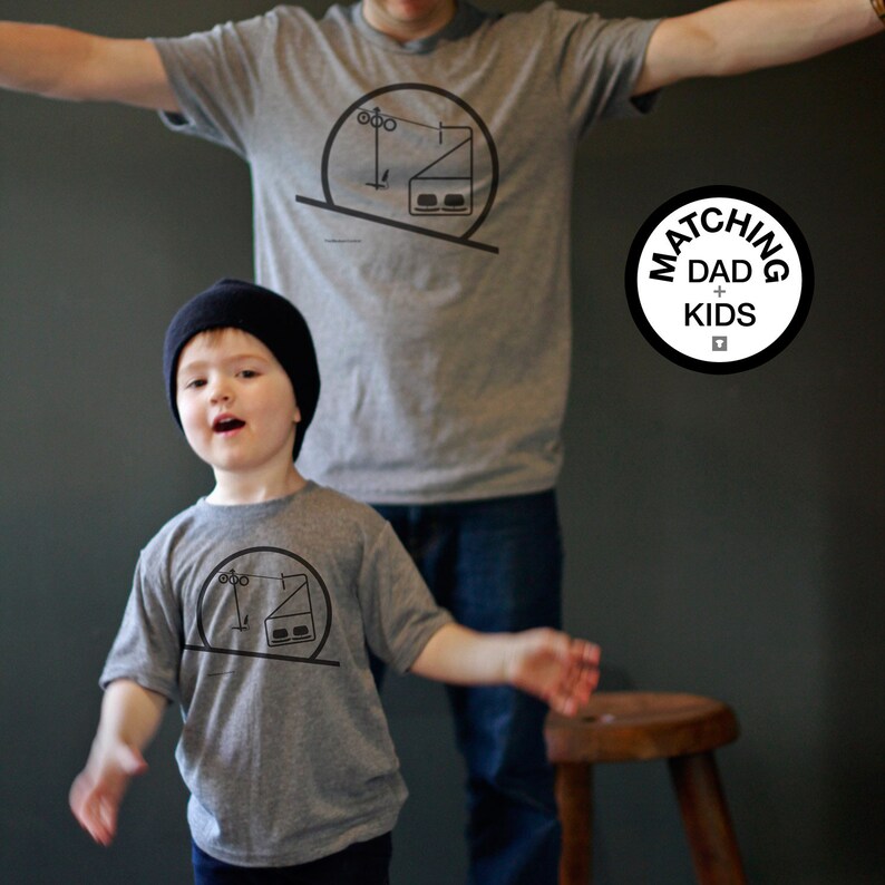 May include: Two grey t-shirts featuring a black graphic print of a ski lift chair within a circular frame. The shirts display the same design on the chest. A circular badge on the right side contains the text MATCHING DAD + KIDS.