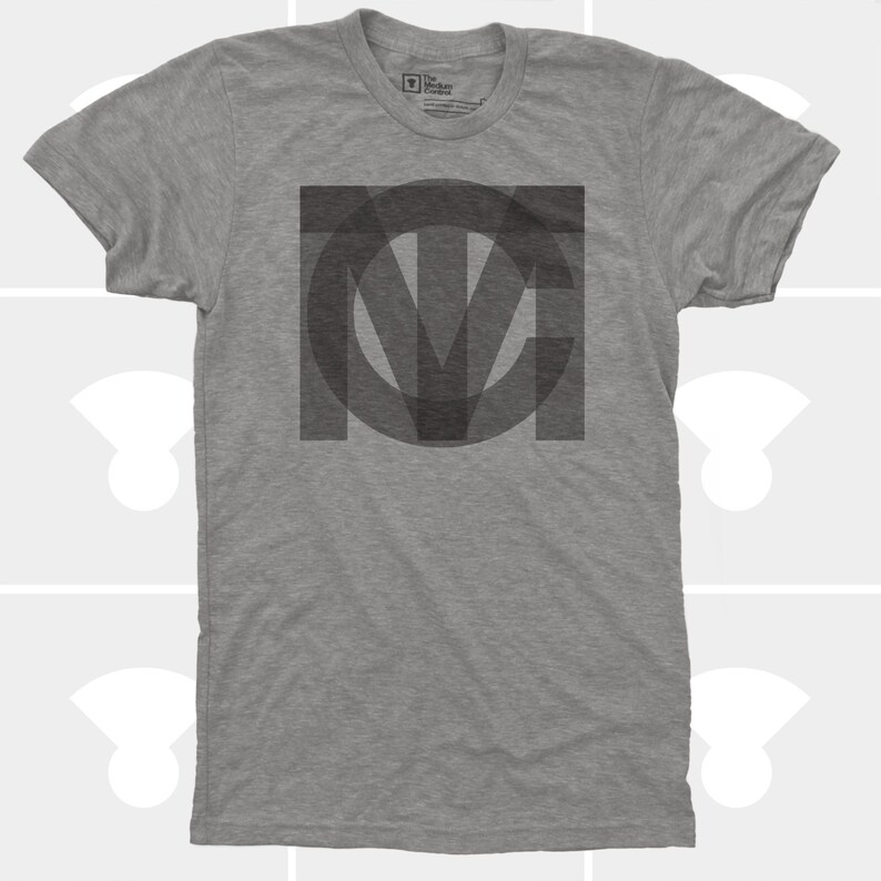 A flat-lay product photo of a heather grey crewneck t-shirt featuring a large, high-contrast interlocking geometric TMC typography logo screen-printed in dark charcoal ink.