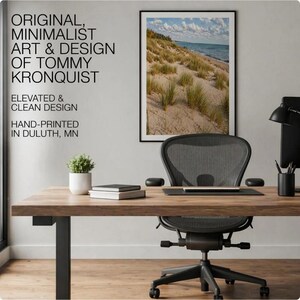 May include: A framed landscape photograph of a sandy beach with dune grass and a blue sky hanging above a wooden desk. The wall features text: ORIGINAL, MINIMALIST ART & DESIGN OF TOMMY KRONQUIST, ELEVATED & CLEAN DESIGN, HAND-PRINTED IN DULUTH, MN.