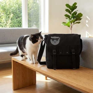 May include: A black messenger bag with a flap closure, two buckles, and a shoulder strap. The bag features a gray graphic of a cat wearing a ninja mask. A black and white cat stands on a wooden bench next to the bag.