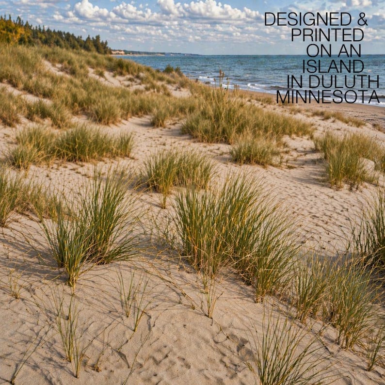 May include: A scenic photograph of a sandy beach with tall green grasses and dunes. The ocean is visible in the background under a blue sky with fluffy white clouds. Text on the top right reads: DESIGNED & PRINTED ON AN ISLAND IN DULUTH MINNESOTA.