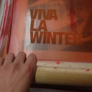 May include: A screen printing process in progress, with the words "VIVA LA WINTER" in orange block letters. A hand holds a wooden squeegee, spreading red ink across the screen.