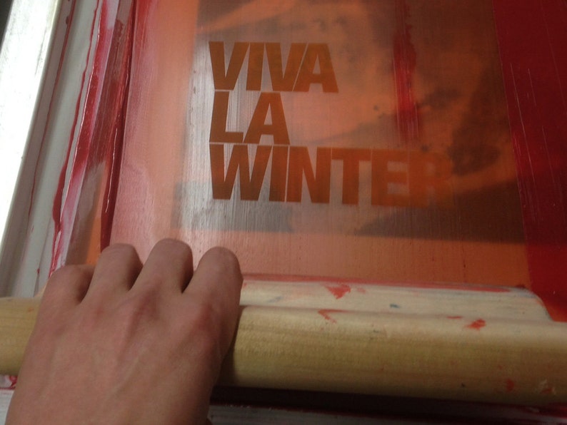 May include: A screen printing process in progress, with the words "VIVA LA WINTER" in orange block letters. A hand holds a wooden squeegee, spreading red ink across the screen.