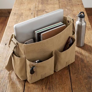 May include: A tan canvas messenger bag with multiple pockets, holding a laptop, notebooks, and a smartphone. A stainless steel water bottle and sunglasses are visible. Keys hang from a pocket. The bag sits on a wooden surface.