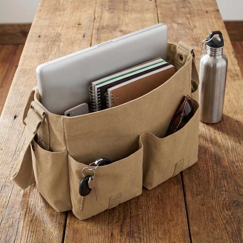 May include: A tan canvas messenger bag with multiple pockets, holding a laptop, notebooks, and a smartphone. A stainless steel water bottle and sunglasses are visible. Keys hang from a pocket. The bag sits on a wooden surface.