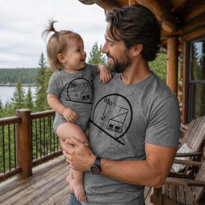 A smiling father holding his baby daughter on a rustic wooden log cabin balcony overlooking a pine forest and lake. Both are wearing matching grey heather tops—the father in a t-shirt and the baby in a short-sleeve bodysuit—featuring a black minimalist graphic of a skiing chairlift layout enclosed in a crisp circle.