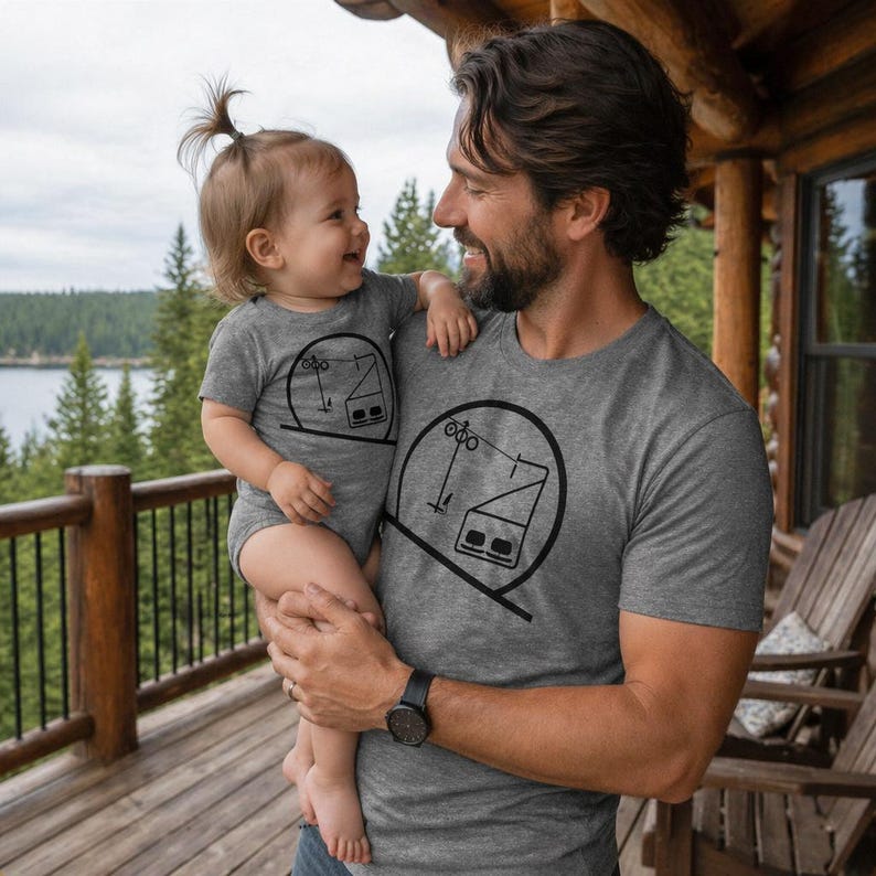 A smiling father holding his baby daughter on a rustic wooden log cabin balcony overlooking a pine forest and lake. Both are wearing matching grey heather tops—the father in a t-shirt and the baby in a short-sleeve bodysuit—featuring a black minimalist graphic of a skiing chairlift layout enclosed in a crisp circle.