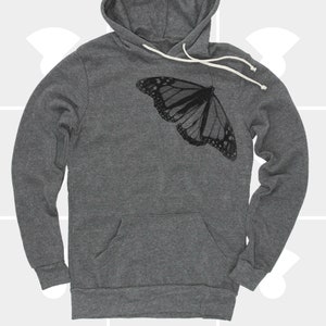 Op de afbeelding: Een donkergrijze hoodie met een zwarte vlinderprint op de voorkant. De vlinder is gecentreerd op de borst en kijkt naar rechts.
