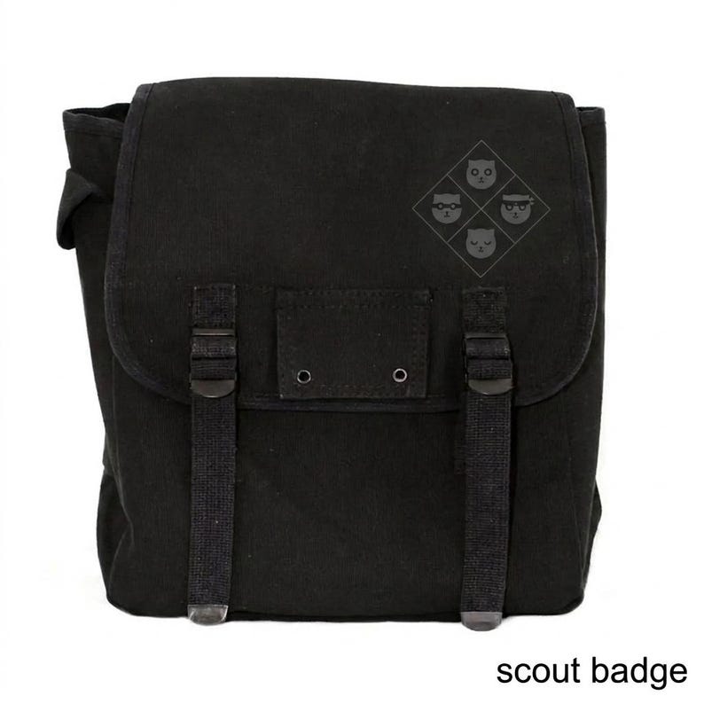 May include: Black canvas messenger bag with a flap closure and adjustable straps. The bag features a small emblem of four cat faces and the text "scout badge" at the bottom.