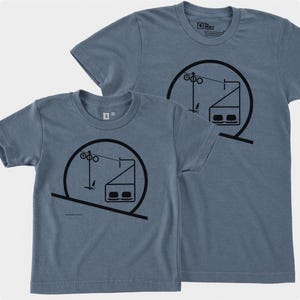 May include: Two heathered blue short-sleeve t-shirts featuring a black graphic print of a ski lift chair within a circular frame. The design includes a cable, support tower, and a two-person chair. The shirts have a crew neckline and a small size tag.
