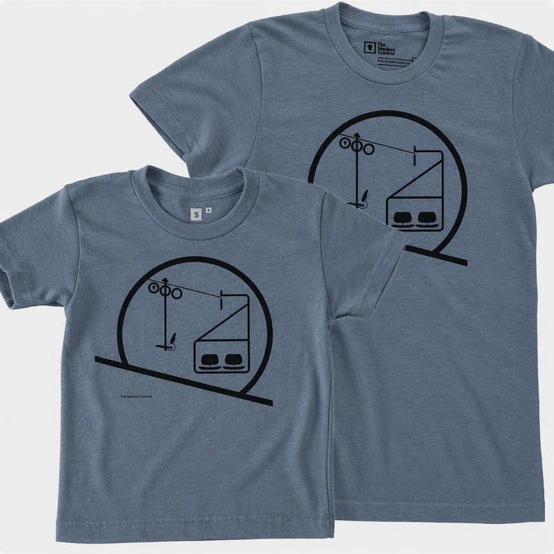 May include: Two heathered blue short-sleeve t-shirts featuring a black graphic print of a ski lift chair within a circular frame. The design includes a cable, support tower, and a two-person chair. The shirts have a crew neckline and a small size tag.