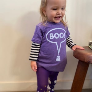 May include: A child wearing a purple Halloween outfit. The outfit includes a purple t-shirt with the word "BOO!" in a speech bubble and a ghost graphic, purple tights, and a black and white striped long sleeve shirt.