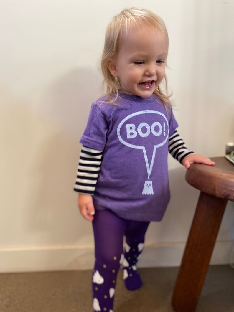 May include: A child wearing a purple Halloween outfit. The outfit includes a purple t-shirt with the word "BOO!" in a speech bubble and a ghost graphic, purple tights, and a black and white striped long sleeve shirt.