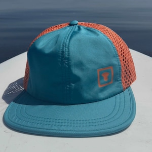 Puede incluir: Una gorra de béisbol de malla turquesa y naranja con un pequeño logotipo cuadrado en la parte delantera.