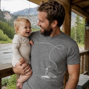 May include: A gray t-shirt with a white graphic of a person fishing on a rock in a lake. A person is holding a baby. The baby is wearing a light-colored onesie. The background shows a river and mountains.