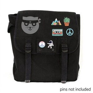 May include: Black canvas backpack with a flap closure, adorned with various enamel pins. Pins include a ninja cat, mountains, a succulent, a cassette tape, a peace sign, and an astronaut. The text "pins not included" is at the bottom.