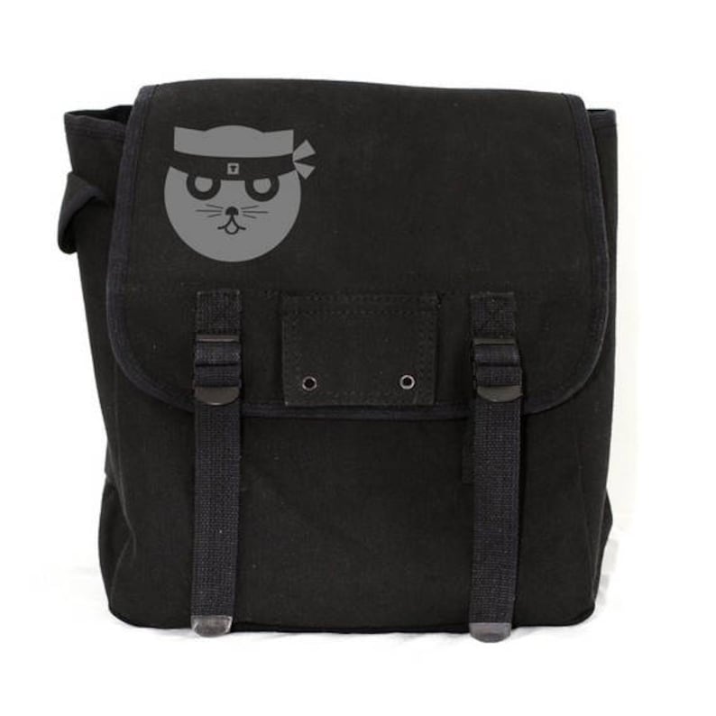 A minimalist black heavyweight canvas backpack featuring a clean, hand-printed graphic of a stylized ninja cat. The pack is displayed in a bright studio setting highlighting its rugged tactical straps and premium textured fabric.
