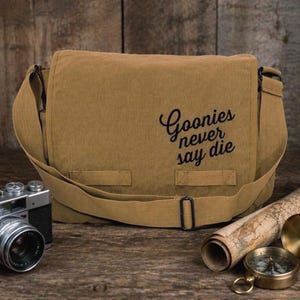 Könnte beinhalten: Eine beige Messenger-Tasche mit dem Text "Goonies never say die" in schwarzer Schrift. Die Tasche hat einen langen Schultergurt und einen Klappenverschluss. Eine Vintage-Kamera, eine gerollte Karte und ein Kompass liegen neben der Tasche.