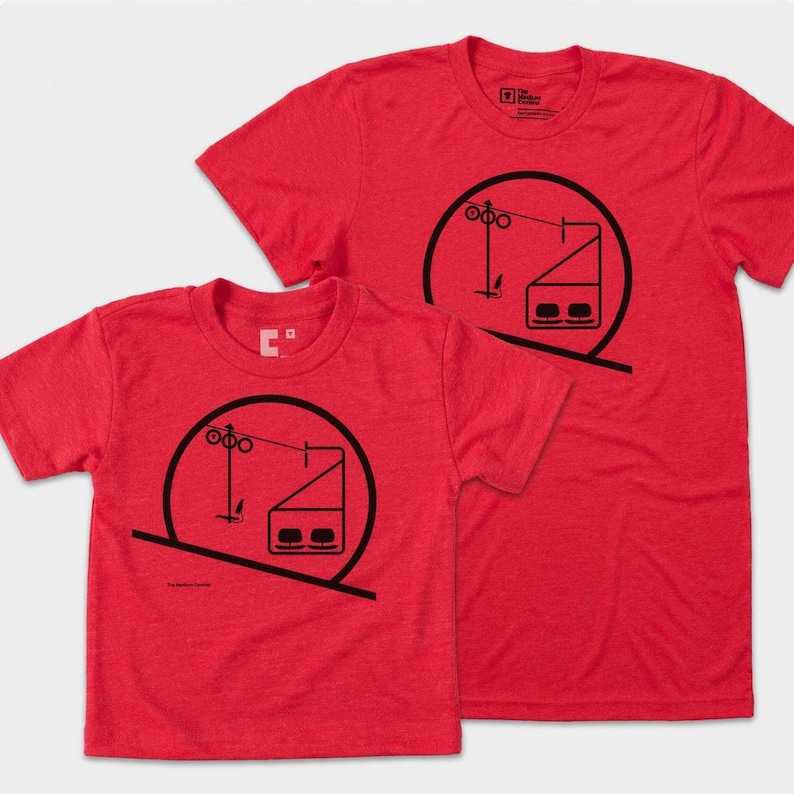 May include: Two red short-sleeved t-shirts featuring a black graphic print of a ski lift chair suspended from a cable within a circular frame. The design includes a stylized pole and a two-seat chairlift carriage, presented on a heathered red fabric.
