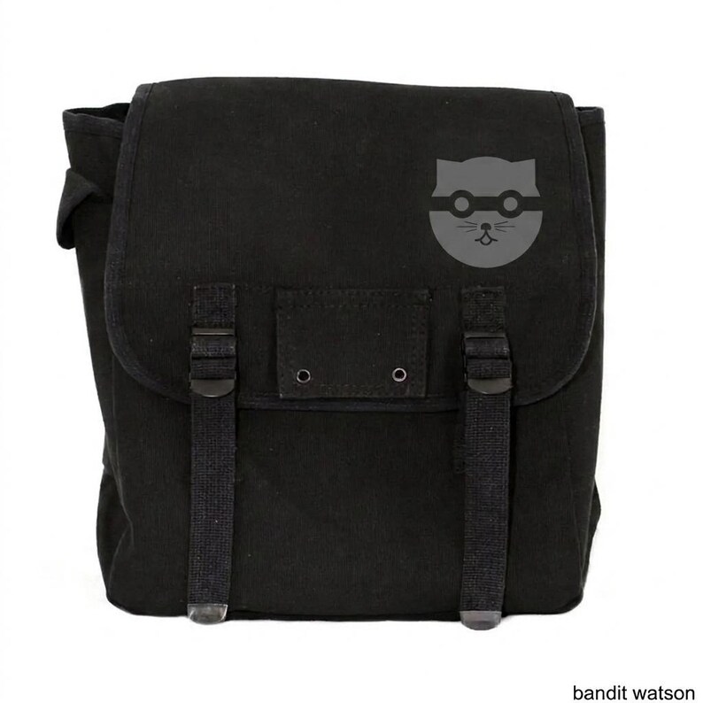 May include: Black messenger bag with a flap closure and adjustable straps. The bag features a gray graphic of a cat wearing glasses. The bag has a rectangular shape and is made of a durable fabric.