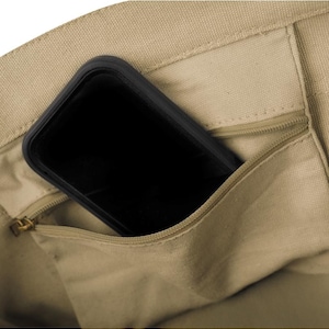 May include: A close-up of a beige canvas tote bag with a black smartphone in the zippered pocket. The zipper is gold-colored. The bag's material is a light tan color.