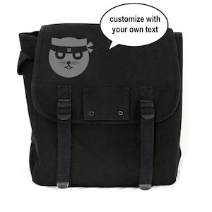 May include: Black canvas backpack with a flap closure and adjustable straps. A gray cat ninja graphic is on the front. The text bubble says "customize with your own text".