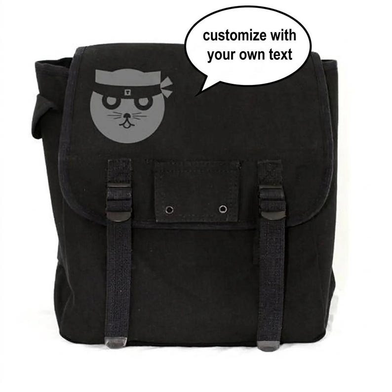May include: Black canvas backpack with a flap closure and adjustable straps. A gray cat ninja graphic is on the front. The text bubble says "customize with your own text".