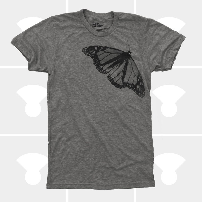 Op de afbeelding: Een heidekleurig T-shirt met een zwart-witte vlinderprint op de voorkant. De vlinder is gecentreerd aan de linkerkant van het shirt.