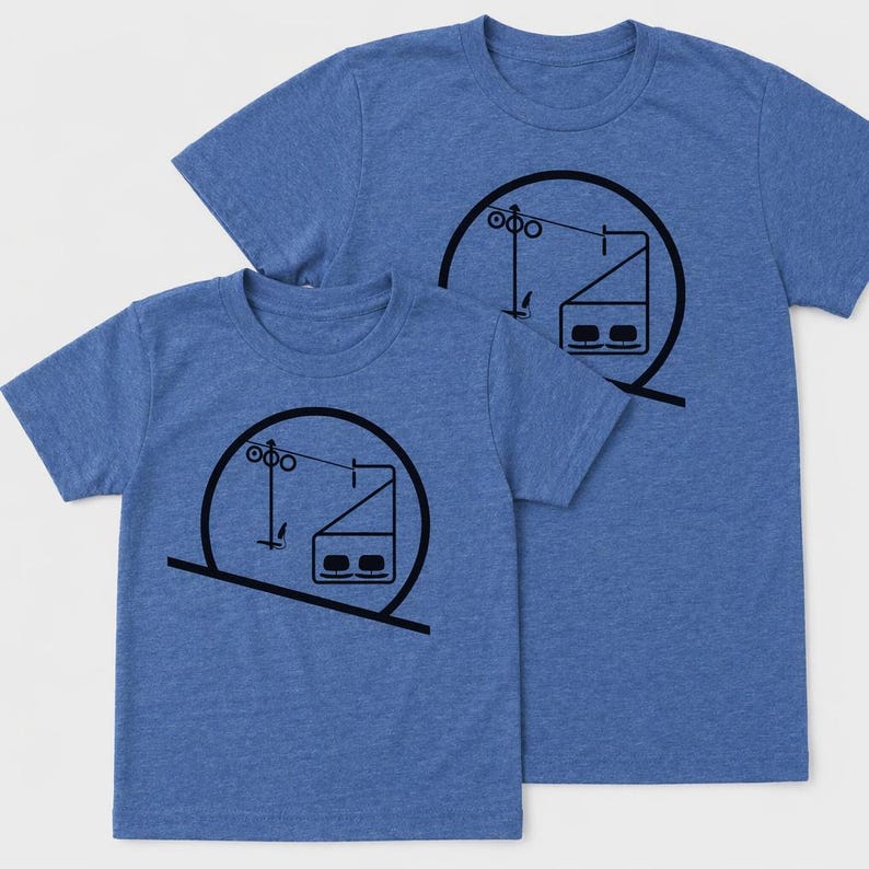 May include: Two blue heathered t-shirts featuring a black graphic print of a ski lift chair within a circular frame. The design includes a stylized cable, support tower, and a two-person chairlift seat, presented in a simple line art style.