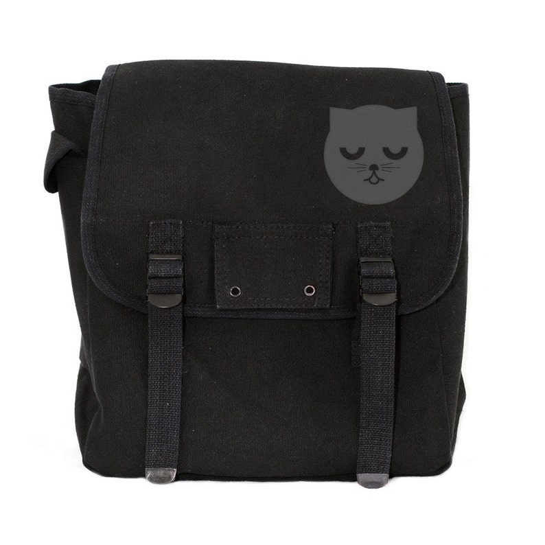 May include: Black canvas messenger bag with a flap closure and adjustable straps. Features a gray cat face graphic on the front. The bag has two buckle closures and a small pocket. The bag is designed to be carried over the shoulder.